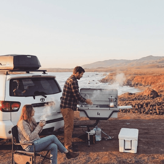 Portable outdoor grill with side shelves and lid, set up on a hitch for cooking