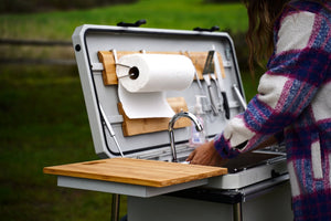 Camp kitchen grill with open lid and cutting board, sink and utensil storage