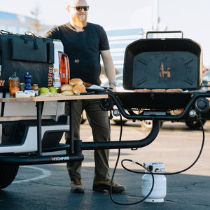 Hitch mounted propane grill with open lid and dual side shelves, dark finish with knobs.