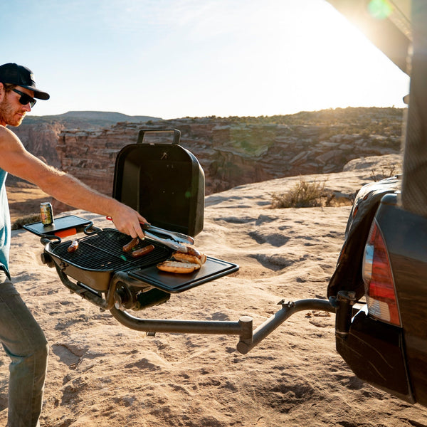 Hitch-mounted propane grill with open lid and cooking grates, man preparing food