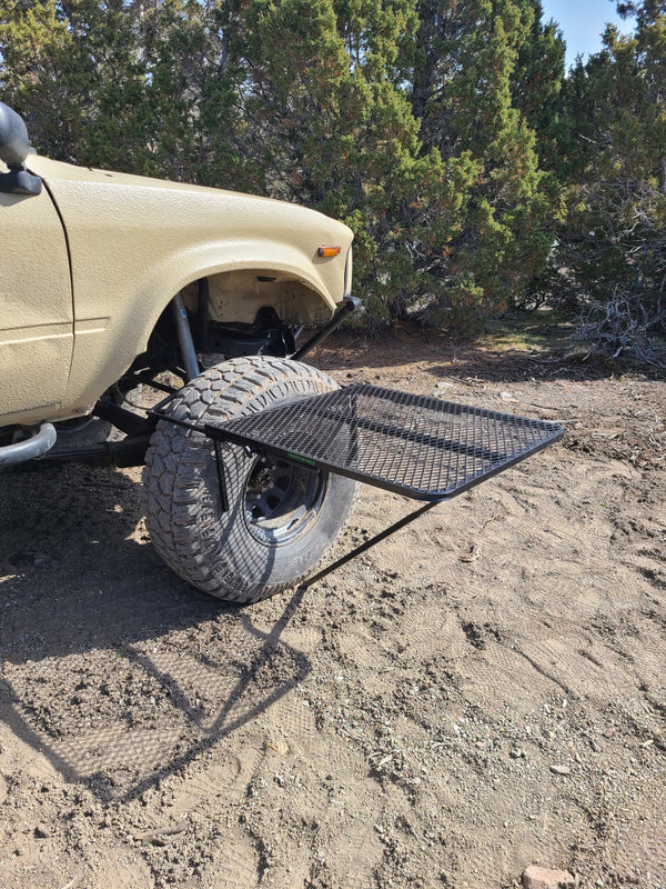 Large black aluminum camping table with wire mesh top on ground near a parked vehicle