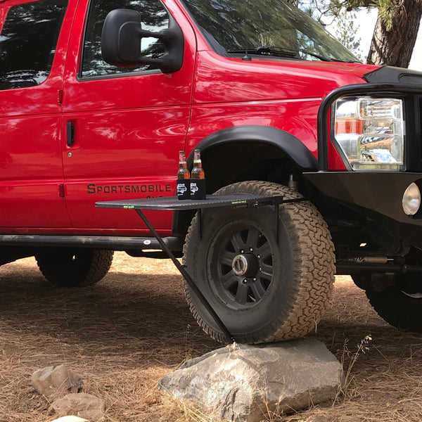 Large black aluminum camping table with wire mesh top on ground near a red truck