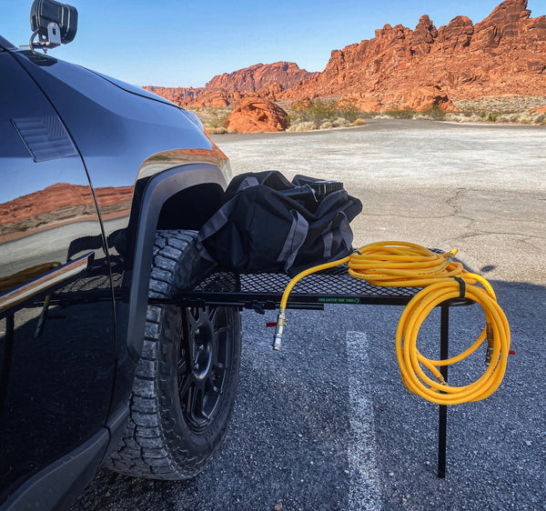 Large black aluminum camping table with wire mesh top attached at the side of a vehicle