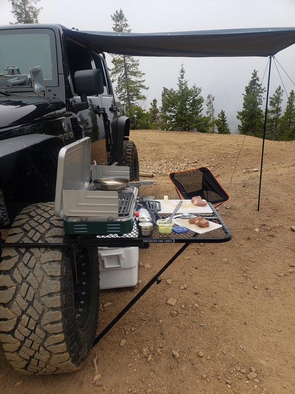 Large black aluminum camping table with wire mesh top on stand beside a camper