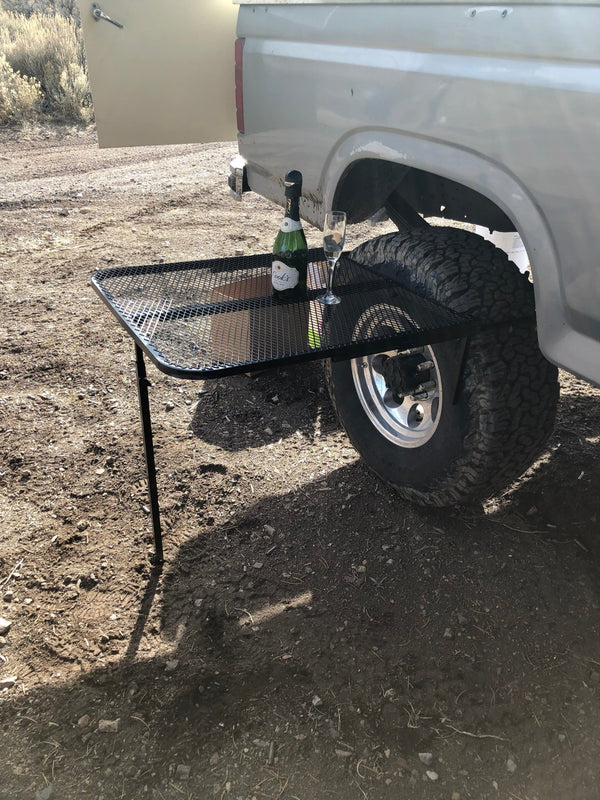 Black steel camping table with wire-mesh top and four legs beside a vehicle