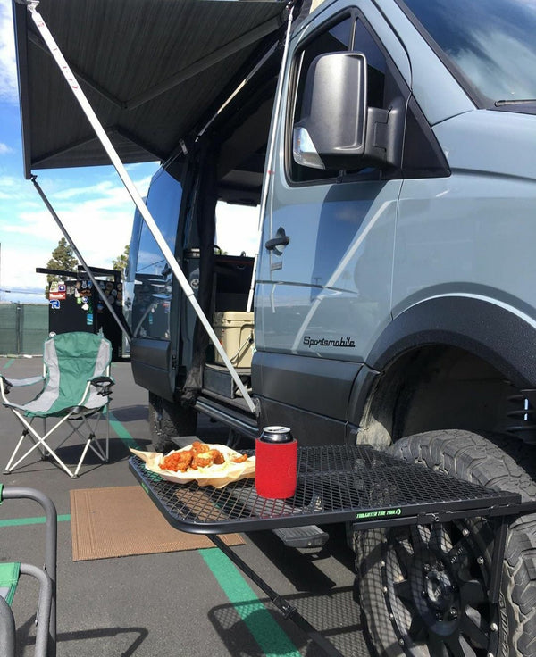 Black steel camping table with rectangular mesh top and folding legs near a van
