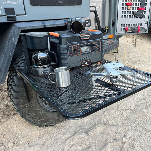 Black aluminum camping table with mesh top and four legs, shown next to a vehicle.