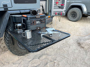 Black aluminum camping tire table with wire mesh top, mug, and accessories on it