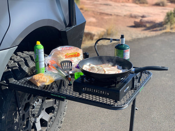 Black wire mesh camping table with raised edge, holding a skillet and bottles.