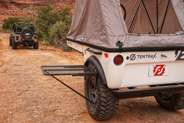 Camping tire table with black metal mesh top and foldable legs, mounted near a trailer.